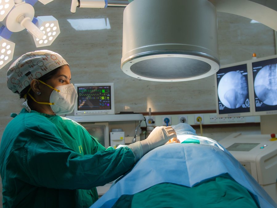 Dr. Kalpana K working under the C-arm with live fluoroscopy images of the spine on screen